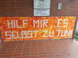 Buntes Schild mit der Aufschrift 'HILF MIR, ES SELBST ZU TUN!' vor einer Backsteinwand